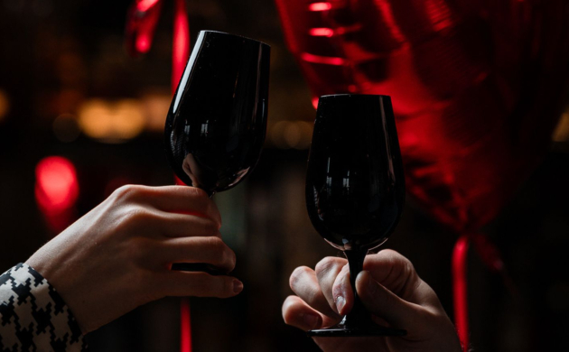 Deux mains trinquant avec un verre de vin devant des ballons en forme de cœur roses, pour une Saint-Valentin romantique.